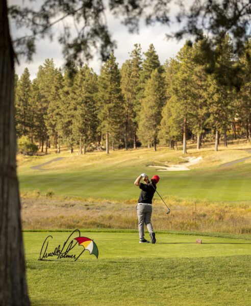 A man swinging a golf club at the tee area, an Arnold Palmer sign with an umbrella sitting on the grass.