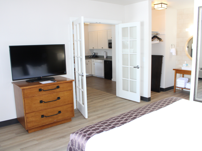 A king-sized bed partially visible with large wide doors that lead into another room. A large TV is seated on a dresser across from the bed.