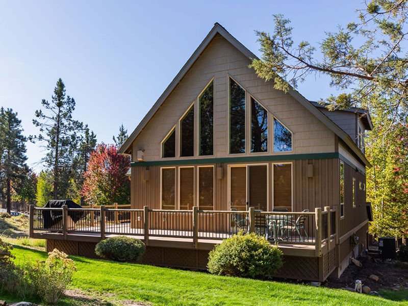 An exterior view of a wooden chalet with large windows and a patio that wraps around the sides and corner of it. Surrounded by green grass and nature.
