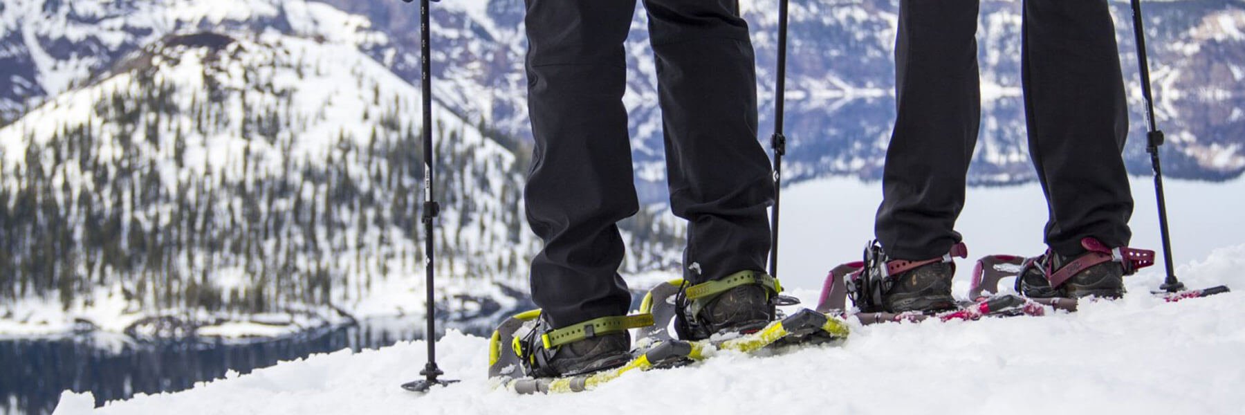 View of two people's legs wearing snowshoes, standing in snow with the snowy mountain set in the background.