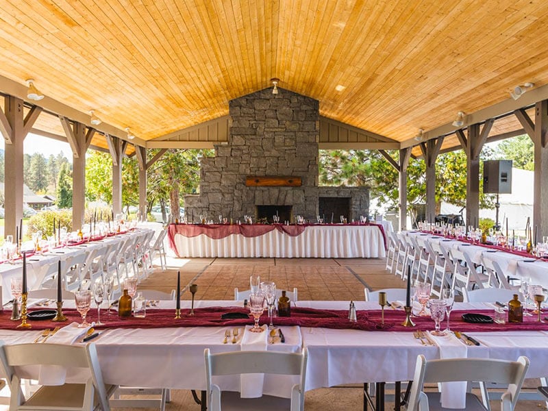A wedding banquet set up inside a wall-less outdoor venue. An arched wooden roof covers the entire space. Tables are adorned with table cloths and red table runners with glassware and candles.
