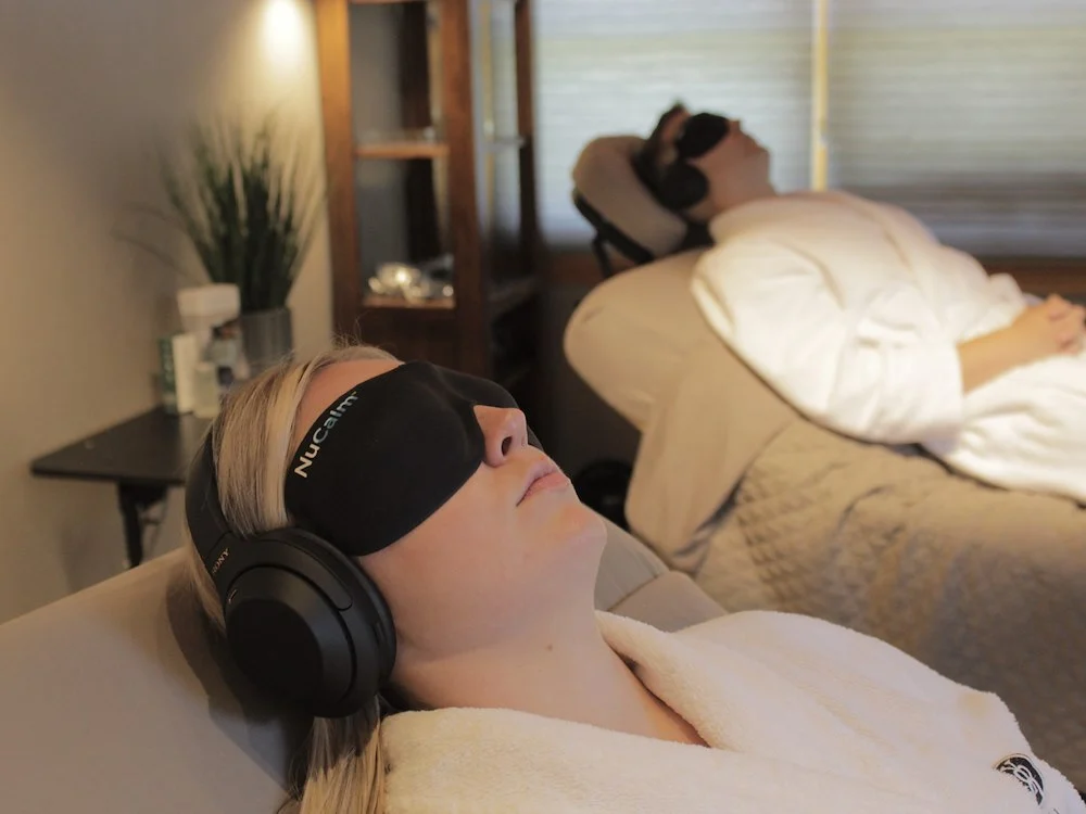 A man and woman with eye masks and headphones wearing robes and laying comfortably on an inclined chair.
