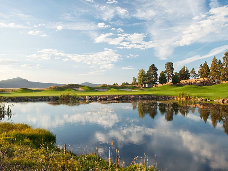 A golf green with a pond set in front of it. Reflections of the golf course, trees, and sky are mirrored in the water.