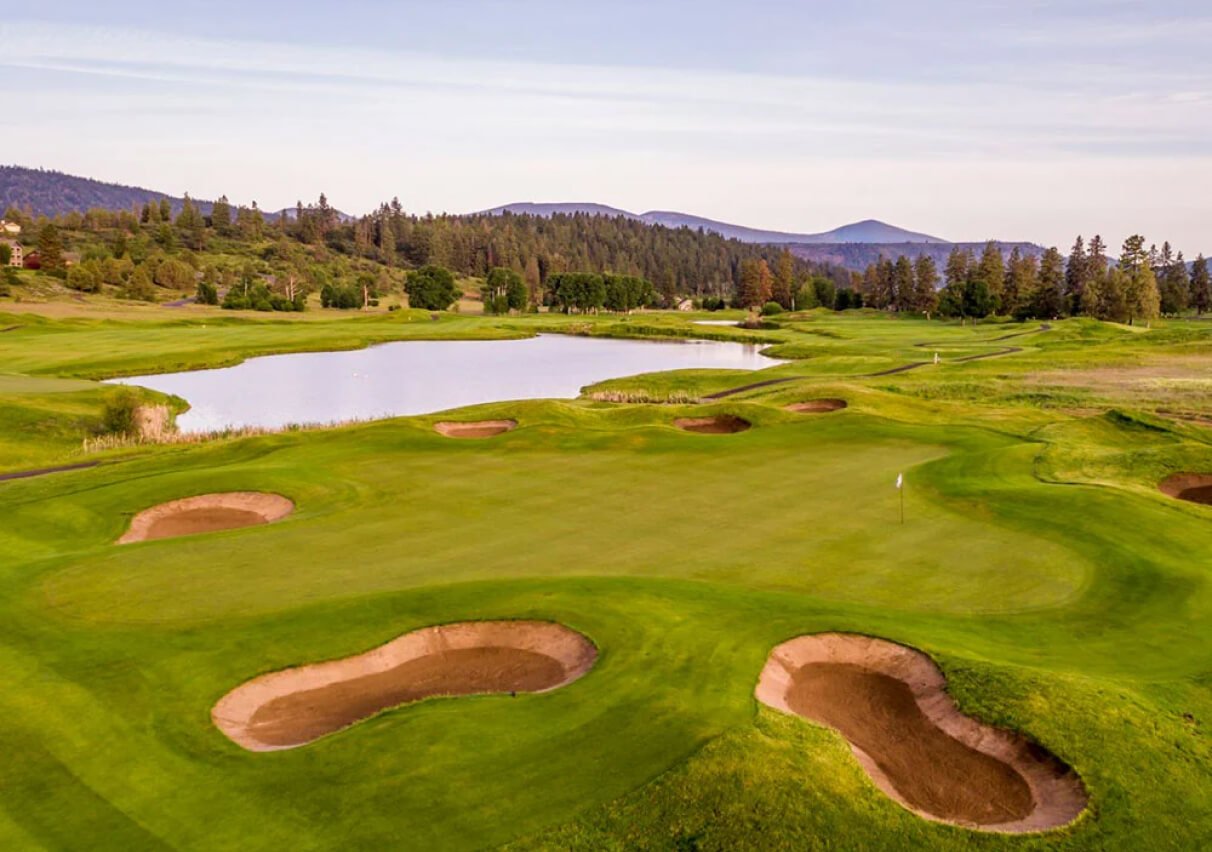 A serene golf course with rolling green fairways, a pristine lake reflecting the clear sky, and majestic mountains in the background under a sunny sky.