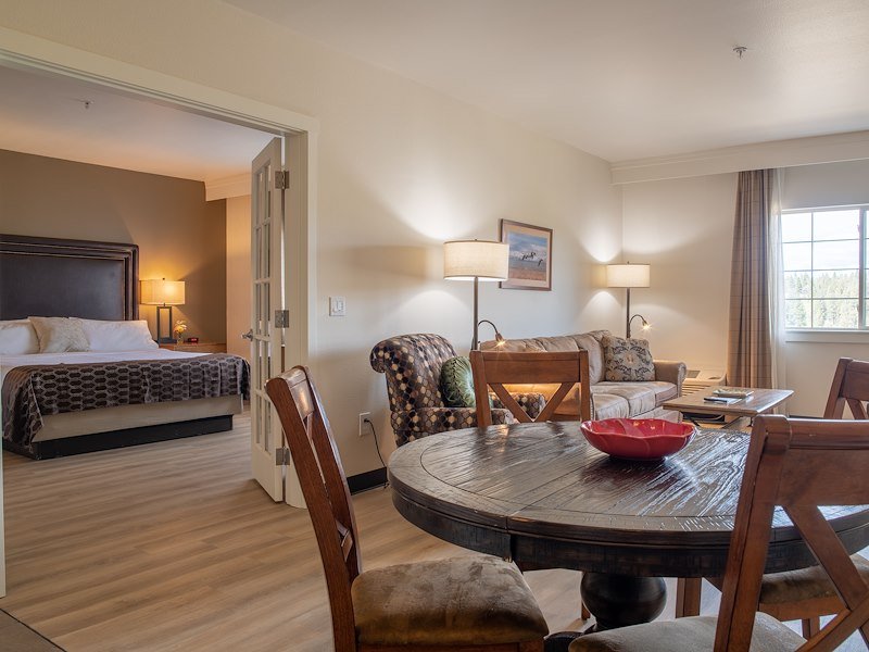 A king-sized bed is visible in the bedroom through open French doors. In the adjoining room, there is a dining table, couch, and coffee table.