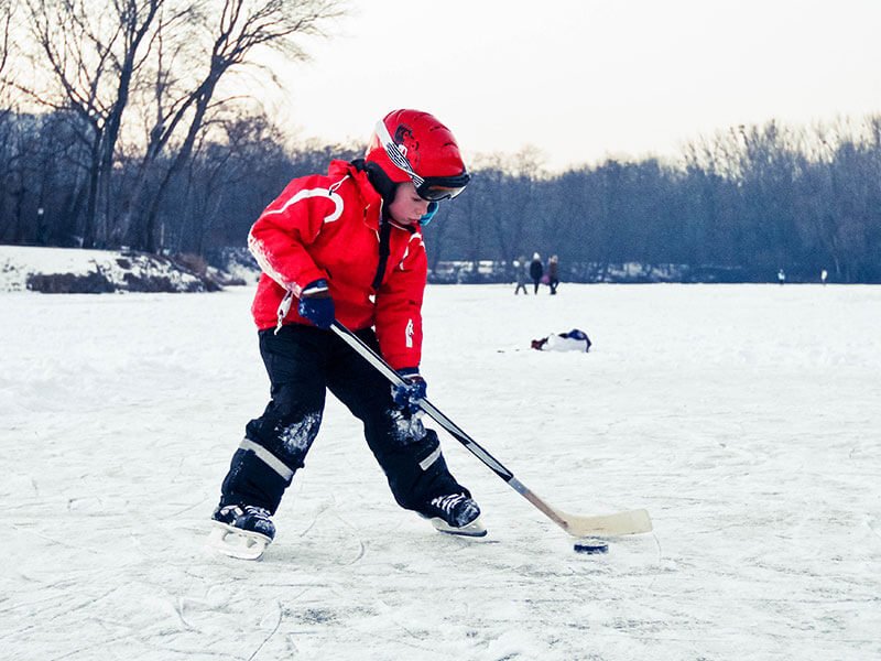 A child dressed in hockey gear and holding a hockey stick with a puck outside on a frozen lake.