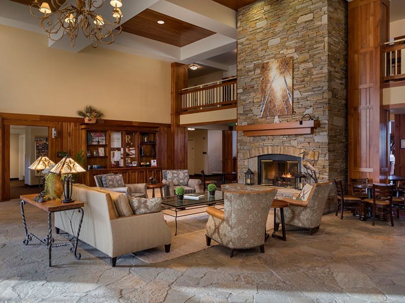A lobby space inside the lodge with 4 large arm chairs and a couch facing towards the center of the room. A large lit fireplace and mantle made of various sized stones is located across from the seating.