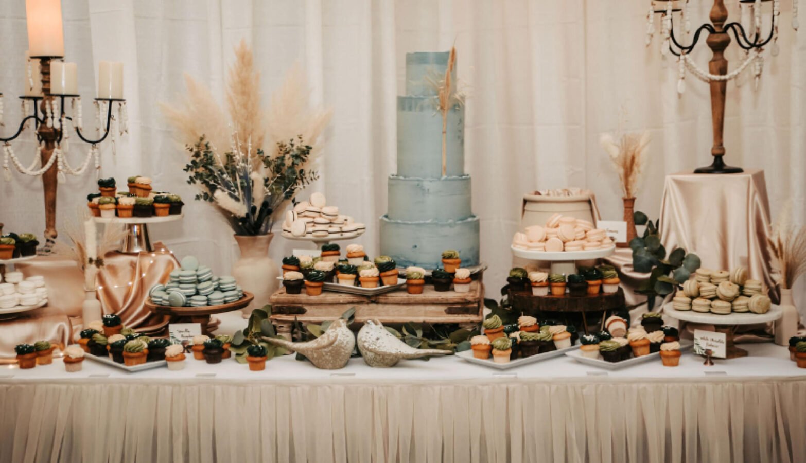 A table full of desserts, macarons and a wedding cake, wedding decorations and candles surround the food.