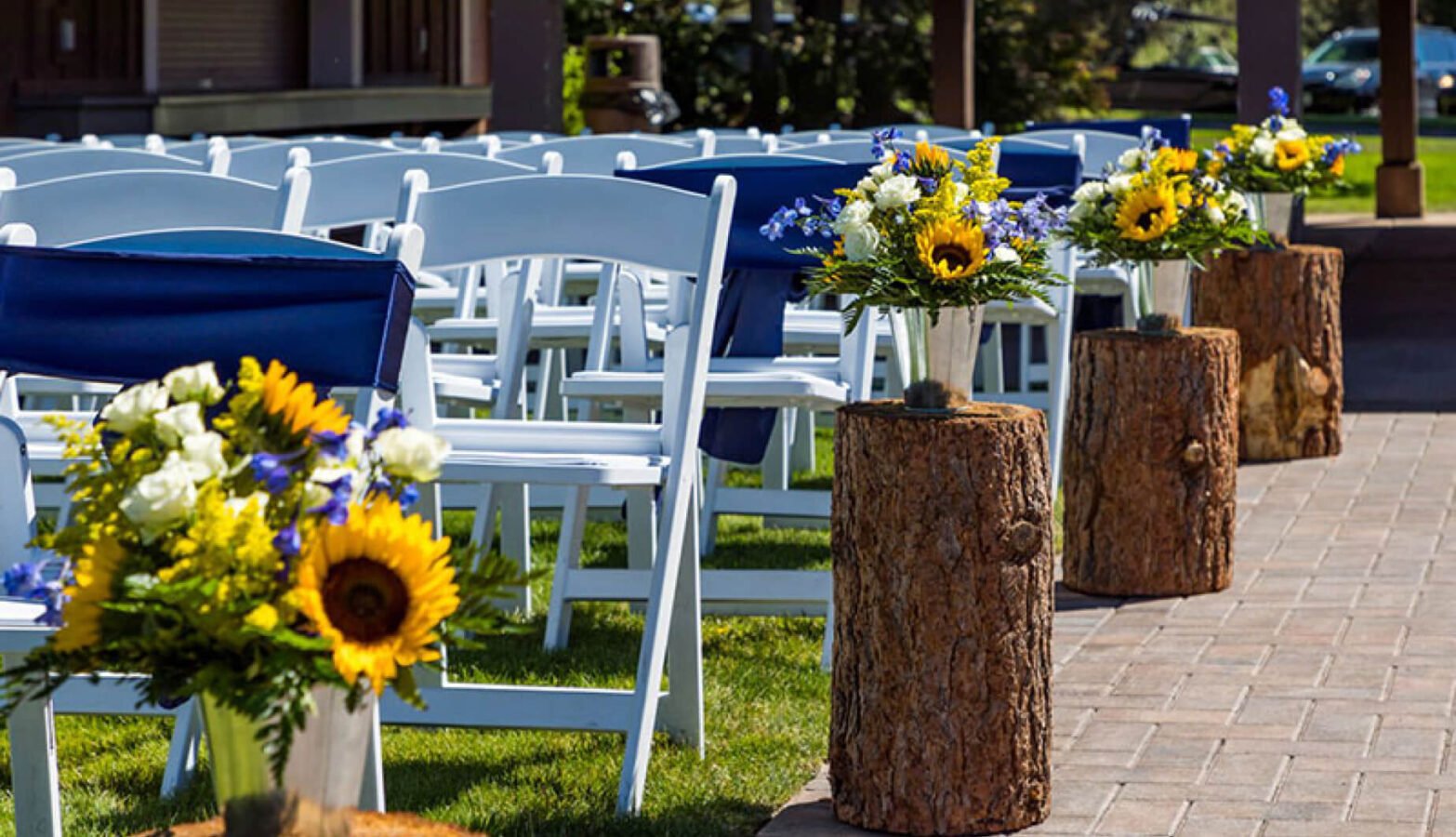 Flower arrangements placed on tree stumps next to rows of white chairs on the lawn.