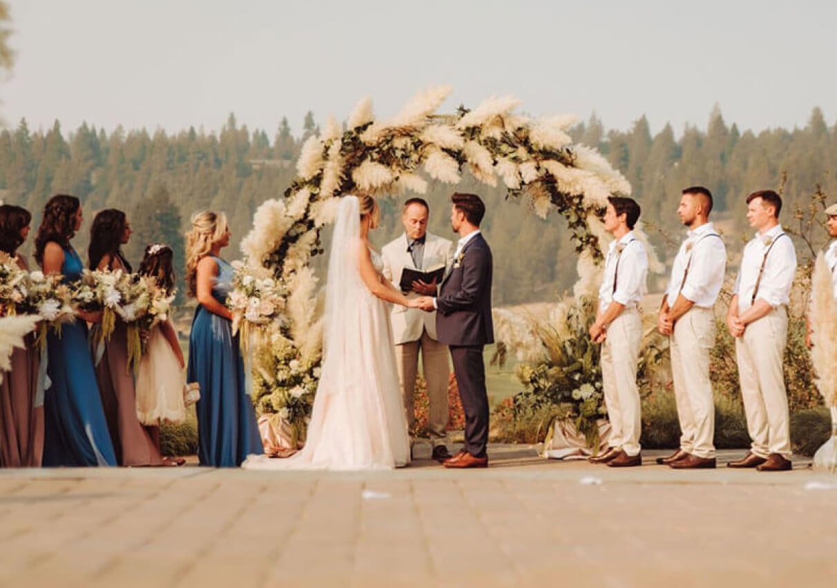 A bride and groom standing in the center with the wedding officiator reading from a book as the two look towards him. The wedding party standing off to the right and left sides, all facing towards the center.