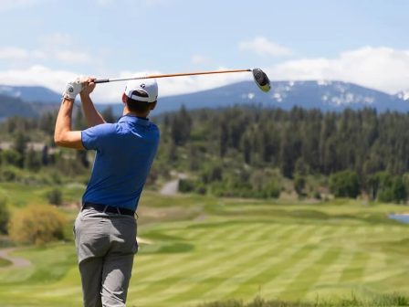 A man swinging a golf club with the golf course pictured behind.
