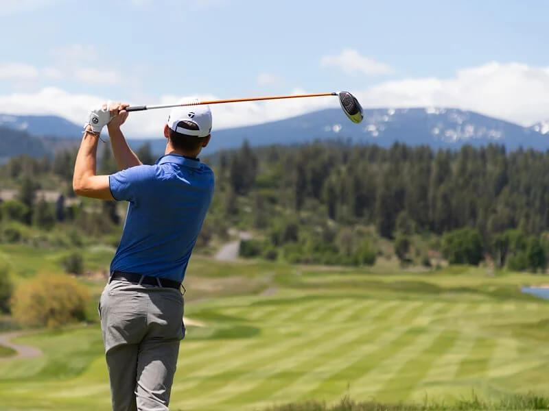 A man swinging a golf club with the golf course pictured behind.