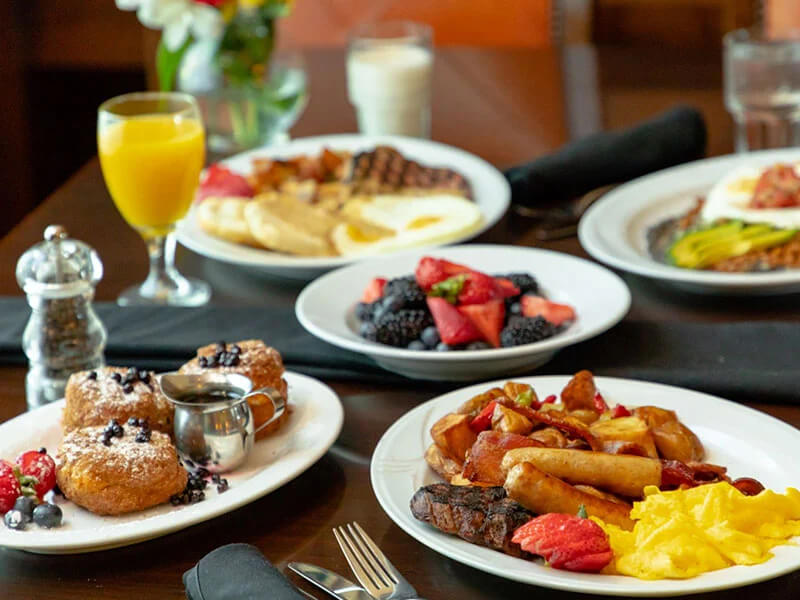 Multiple plates of breakfast items, including pastries, scrambled eggs, and sausage links, accompanied by glasses of orange juice, milk, and water.