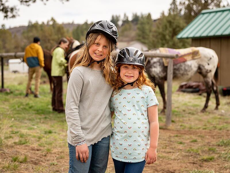 Two young people wearing riding helmets standing next to each other with horses stabled behind them.